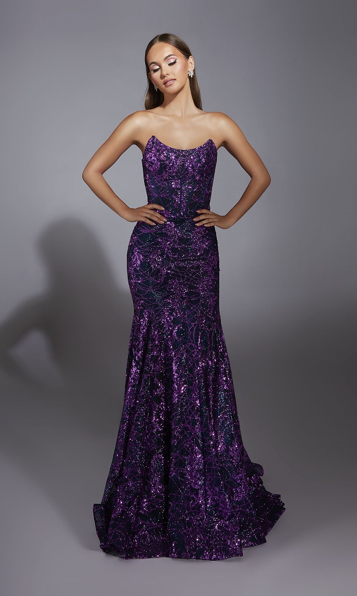 A woman stands on a gray backdrop wearing the Alyce 61887 long prom dress, a strapless mermaid gown in dark purple with shimmering embellishments on the fitted bodice and a flared sequin skirt.