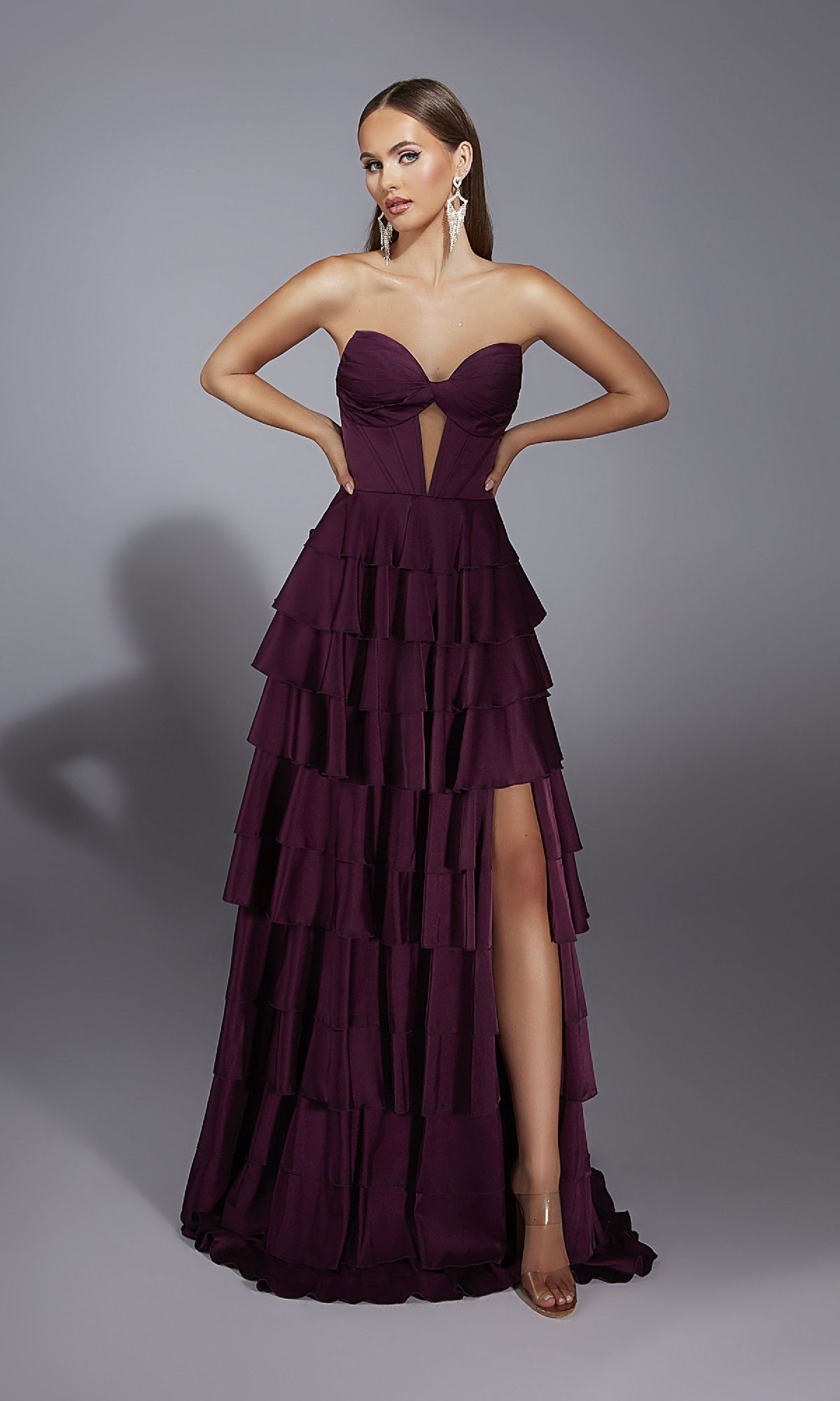 A woman in the Alyce 61889 long prom dress with a strapless sweetheart neckline, high slit, and tiered a-line skirt stands against a gray background, wearing long earrings.
