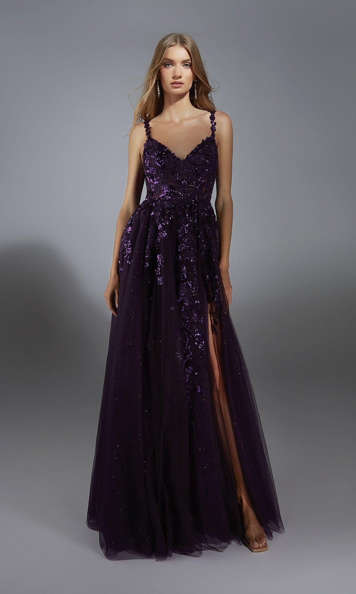 A woman models the Alyce 61891 long prom dress in dark purple, with floral lace appliques and a sheer overlay, plus an elegant A-line glitter tulle skirt, standing before a plain gray background.