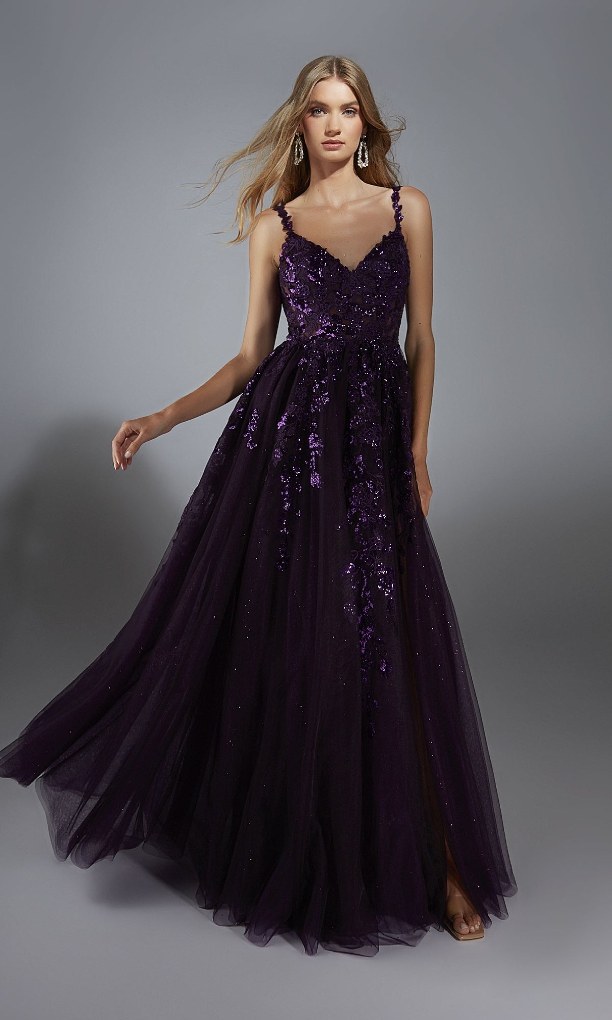 A woman stands against a gray background wearing the Alyce 61891 long prom dress, featuring dark purple sequin embellishments, lace appliques, an elegant sleeveless design, and a floor-length a-line glitter tulle skirt.