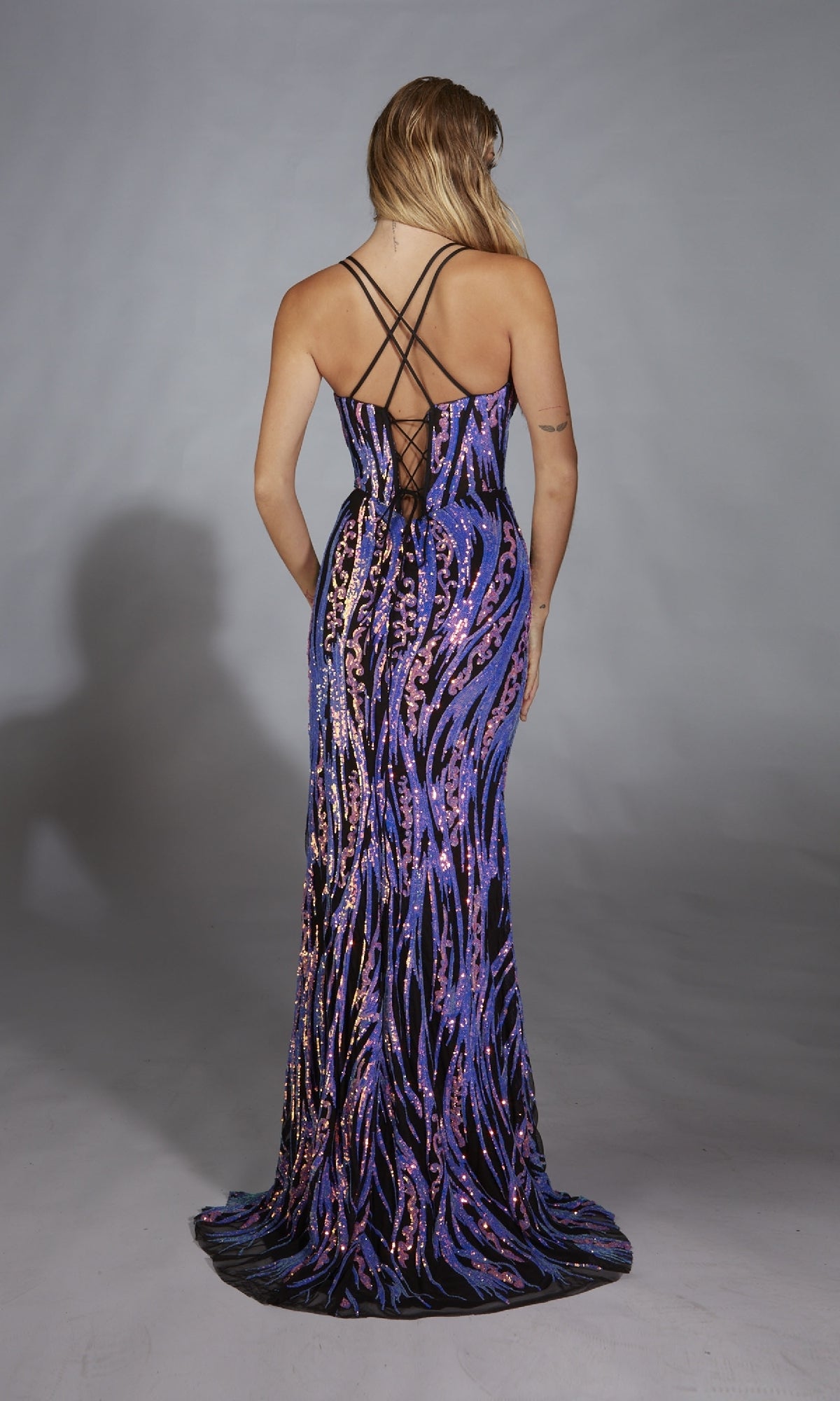 A woman models the Alyce 61897 long prom dress, featuring floor-length purple and gold sequins, crisscross straps, and a corset lace-up back, as she stands facing away from the camera against a gray backdrop.