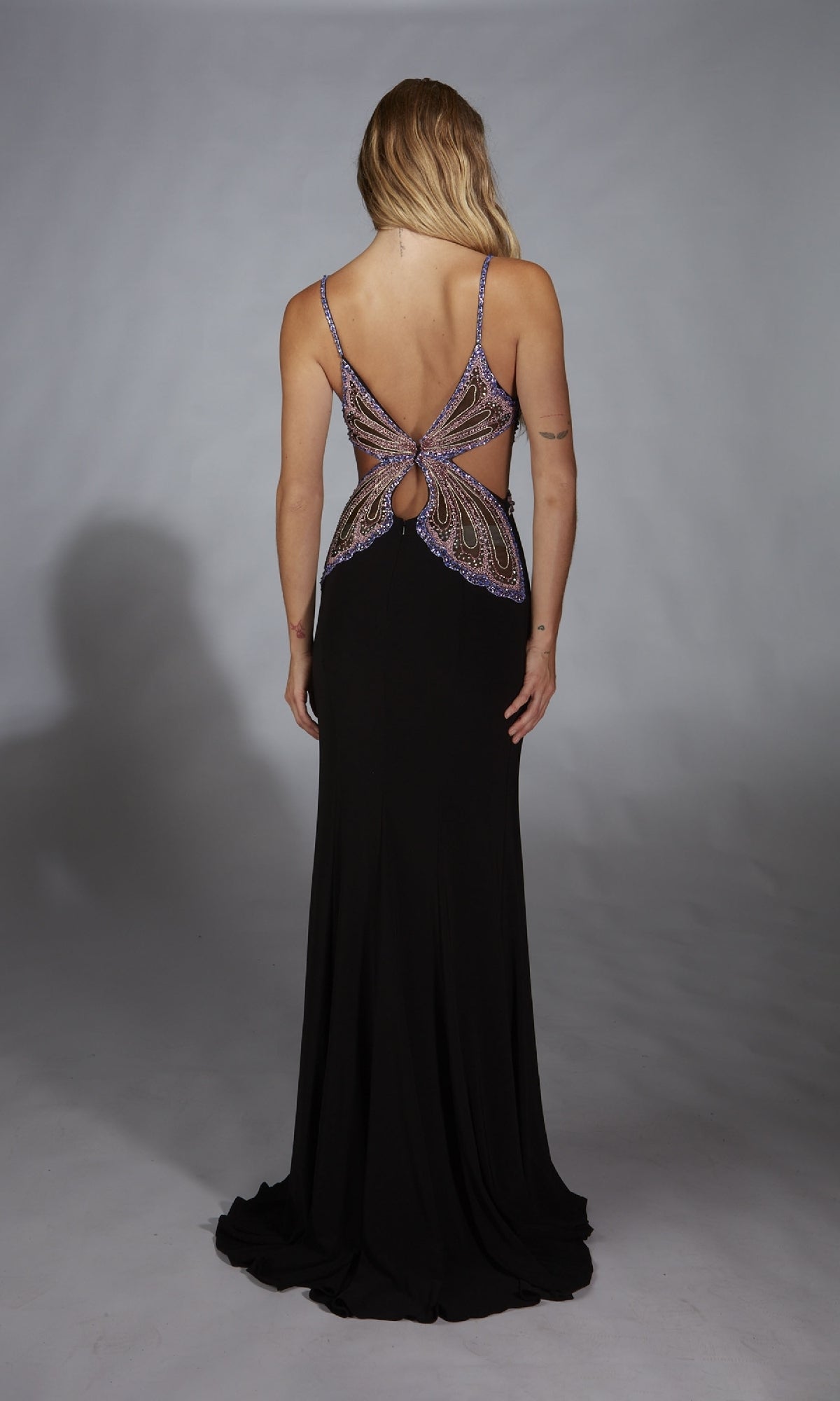 A woman faces away, wearing the Alyce 61902 long prom dress in black with purple and silver accents, a butterfly back cut-out, and a subtle leg slit.