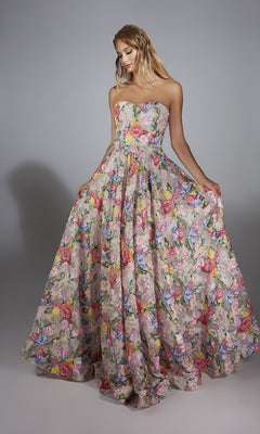 A woman stands against a gray background, holding the sides of her Alyce 61913 long prom dress—a strapless, floor-length gown with a floral print and full skirt.
