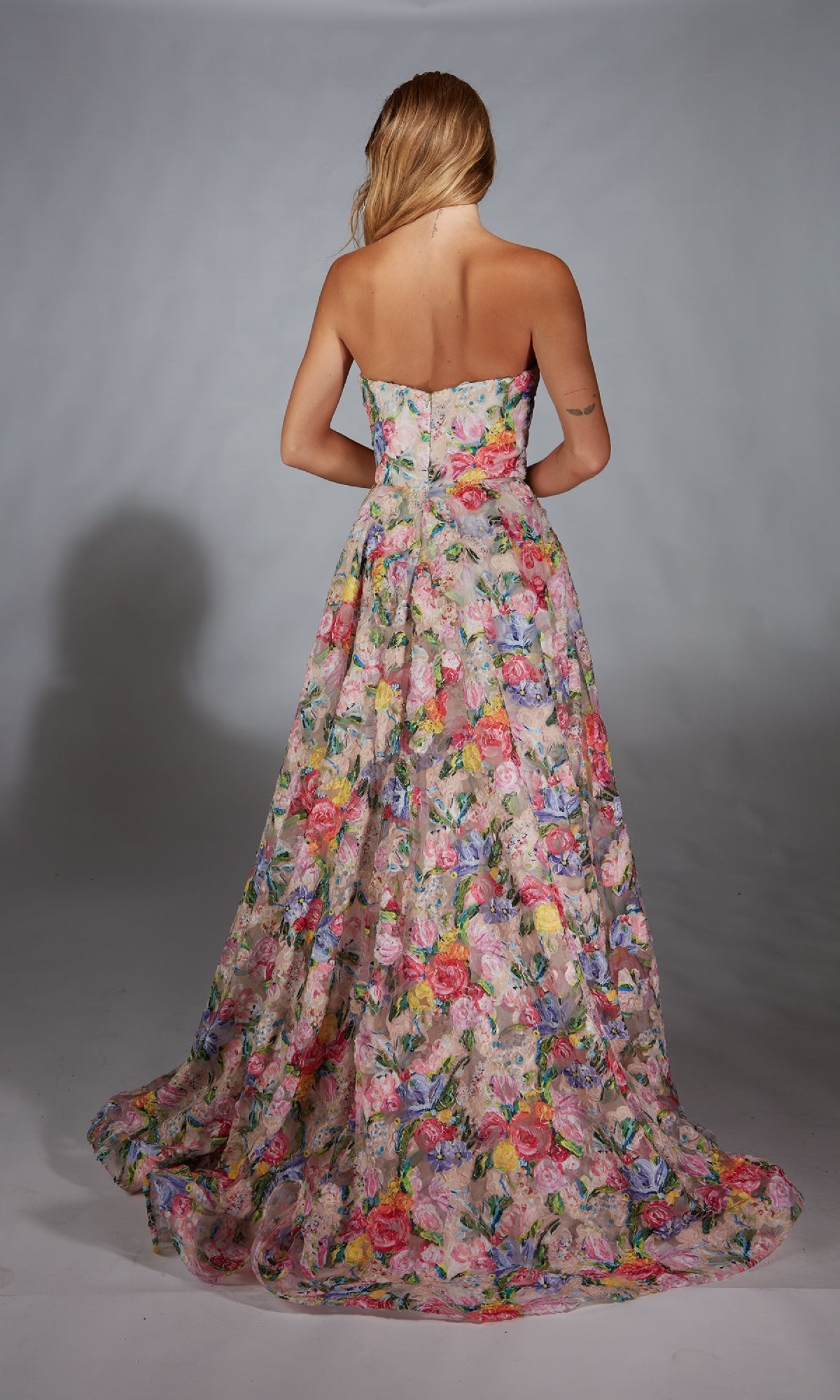 A woman with long blond hair wears the Alyce 61913 Long Prom Dress, a strapless, floor-length floral gown, photographed from behind against a plain gray background.