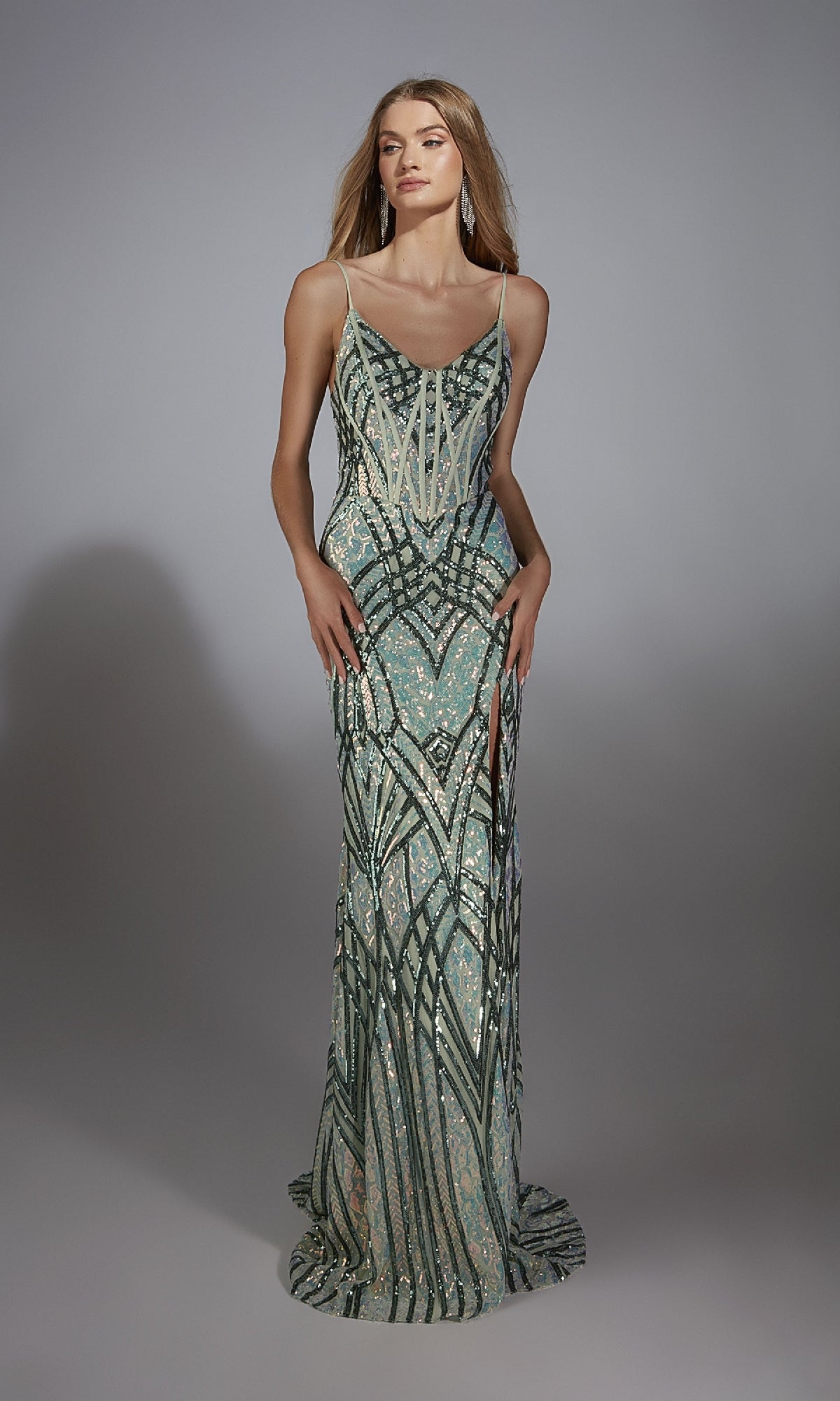 A woman models the Alyce 61920 long prom dress with green, silver, and cream geometric sequins, a corset bodice, high slit, and lace-up back against a plain gray background.