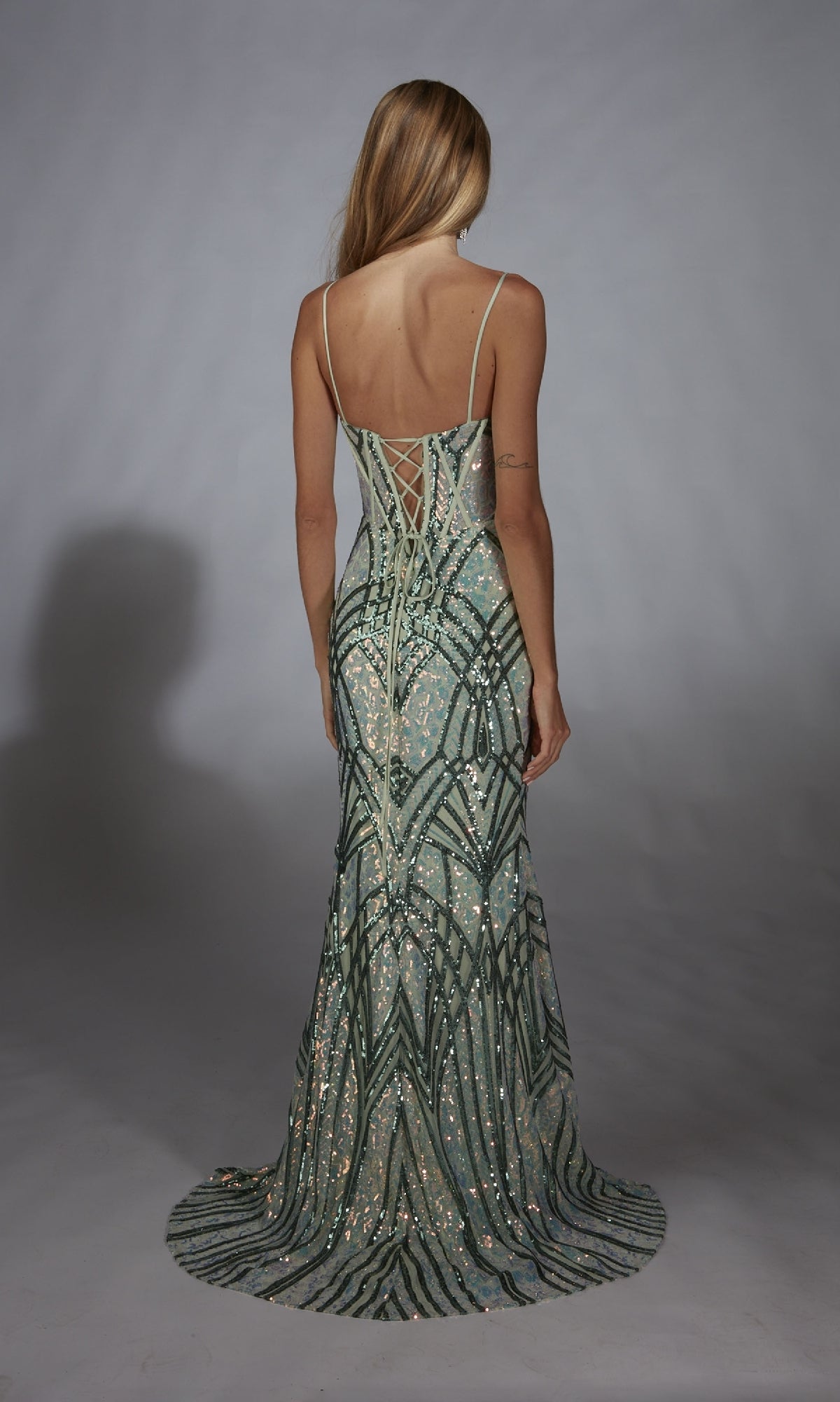 A woman with long blonde hair models the Alyce 61920 long prom dress, a green and silver sequin gown with a lace-up back, standing with her back to the camera against a plain gray background.
