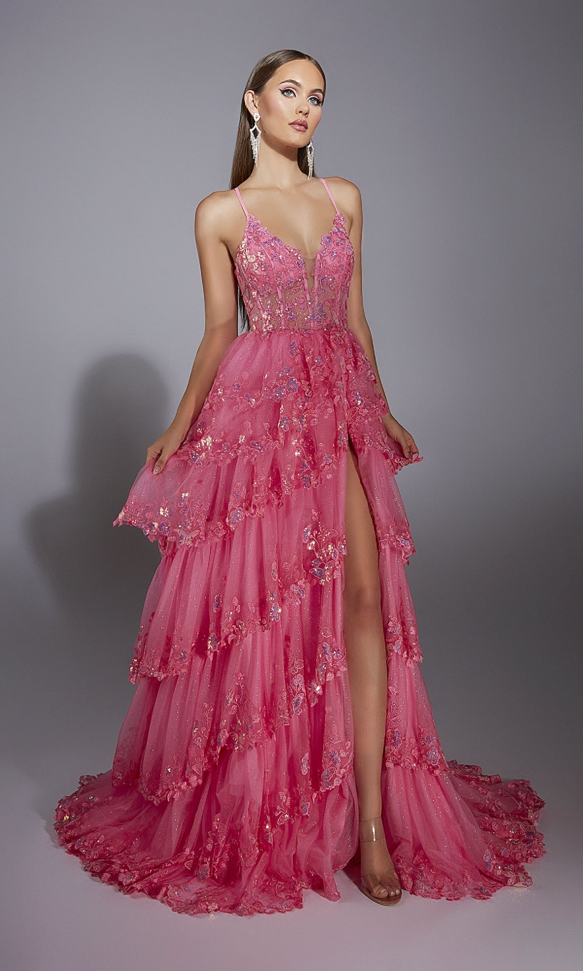 A woman stands against a plain gray background wearing the Alyce 61927 long prom dress: a tiered, bright pink sleeveless gown with a sheer corset bodice, floral embellishments, and a high slit.