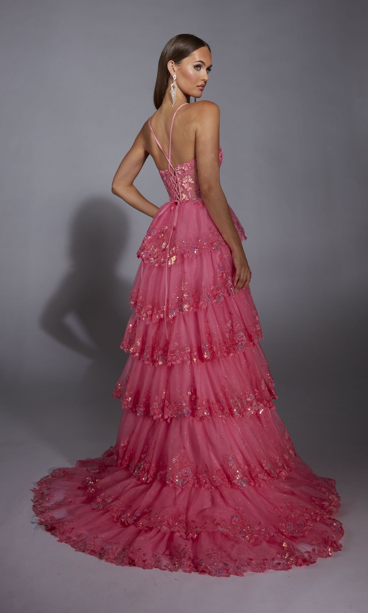 Model poses with her back turned, looking over her shoulder, in the Alyce 61927 Long Prom Dress—a floor-length pink gown featuring tiers, floral embellishments, and a sheer corset bodice—set against a plain gray background.