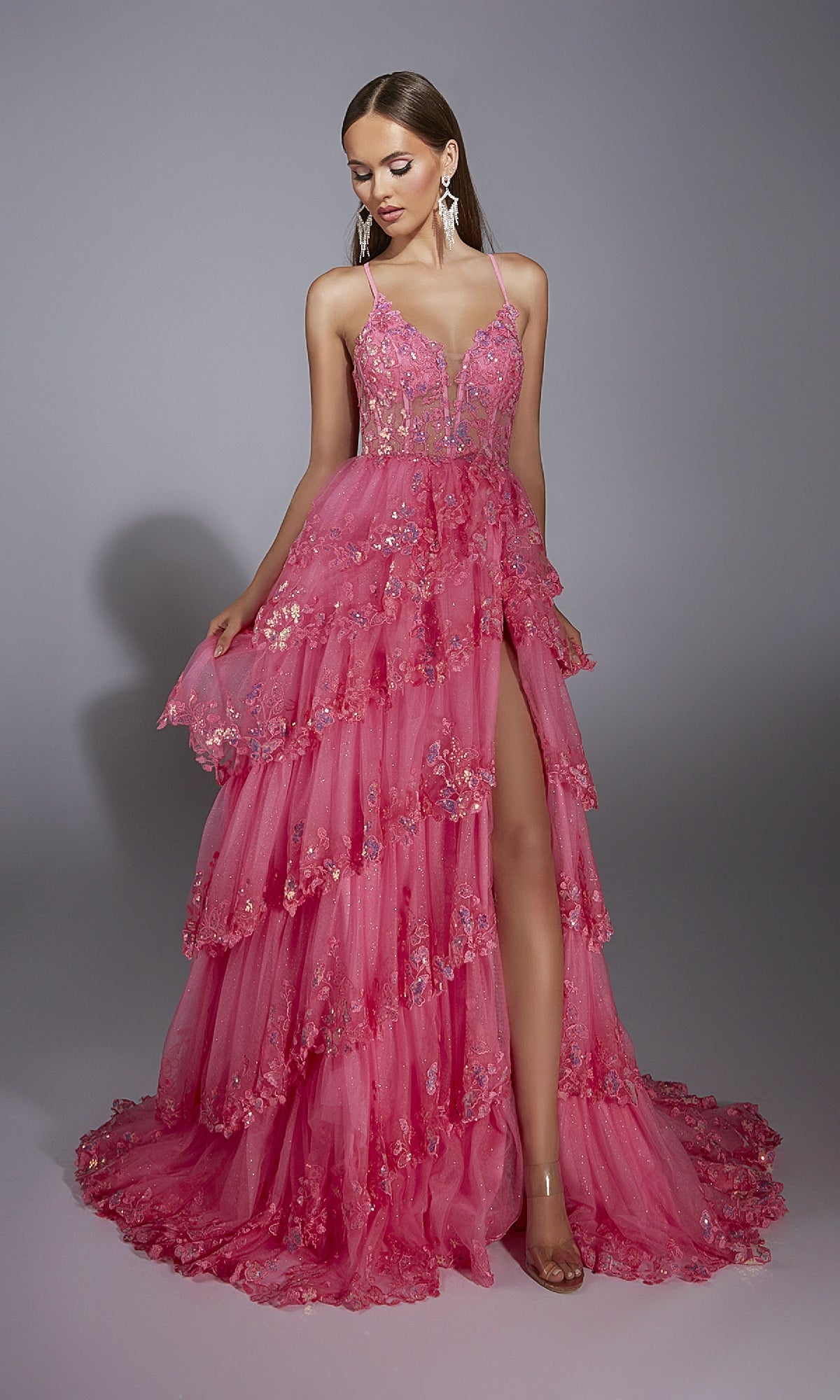A woman stands against a gray background wearing the Alyce 61927 Long Prom Dress, a bright pink tiered tulle gown with sequin lace floral details, deep V-neckline, thin straps, and a high leg slit.