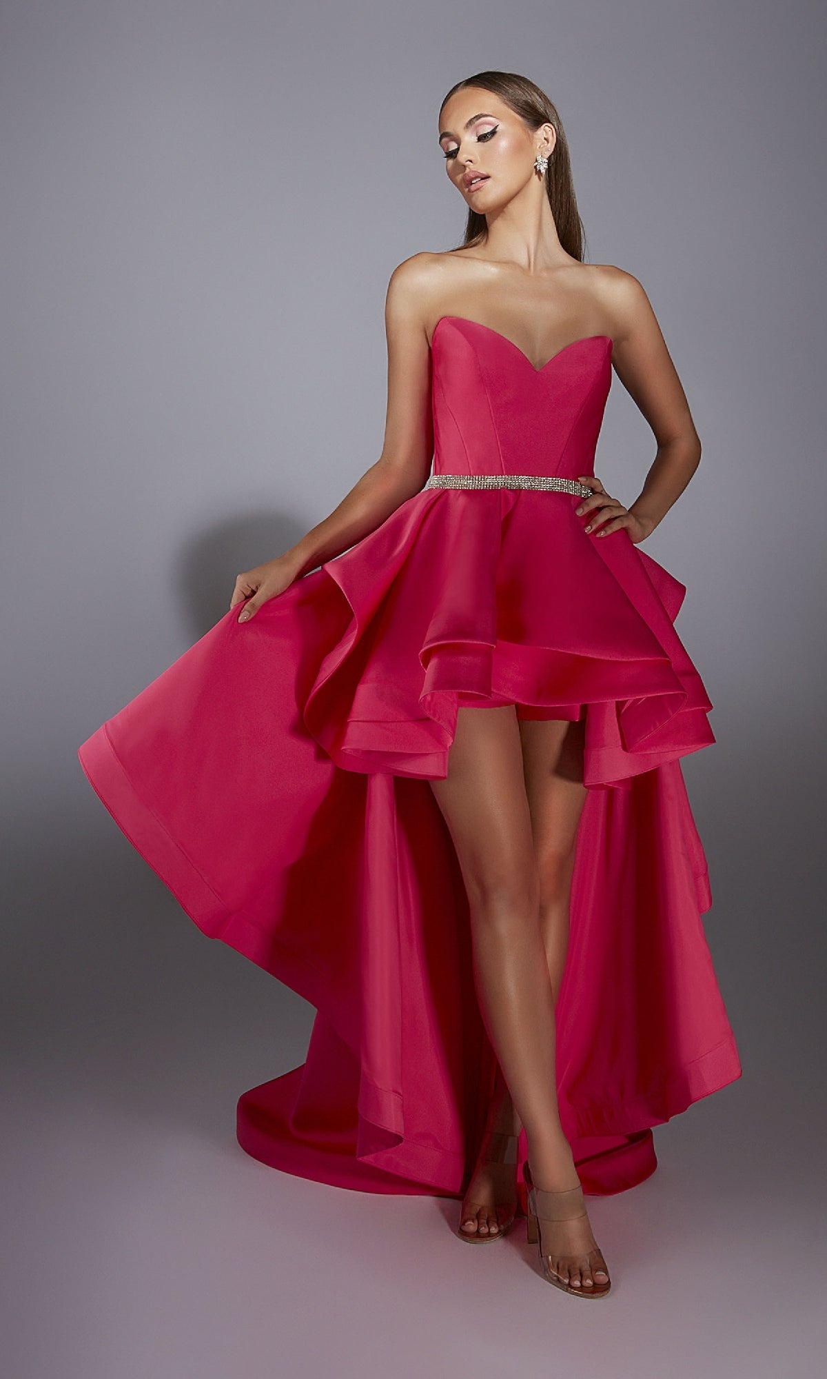 Woman wearing the Alyce 61934 High-Low Strapless Prom Dress in bright pink with a sweetheart neckline, beaded waistline, layered A-line skirt, and transparent high heels, standing against a plain gray background.