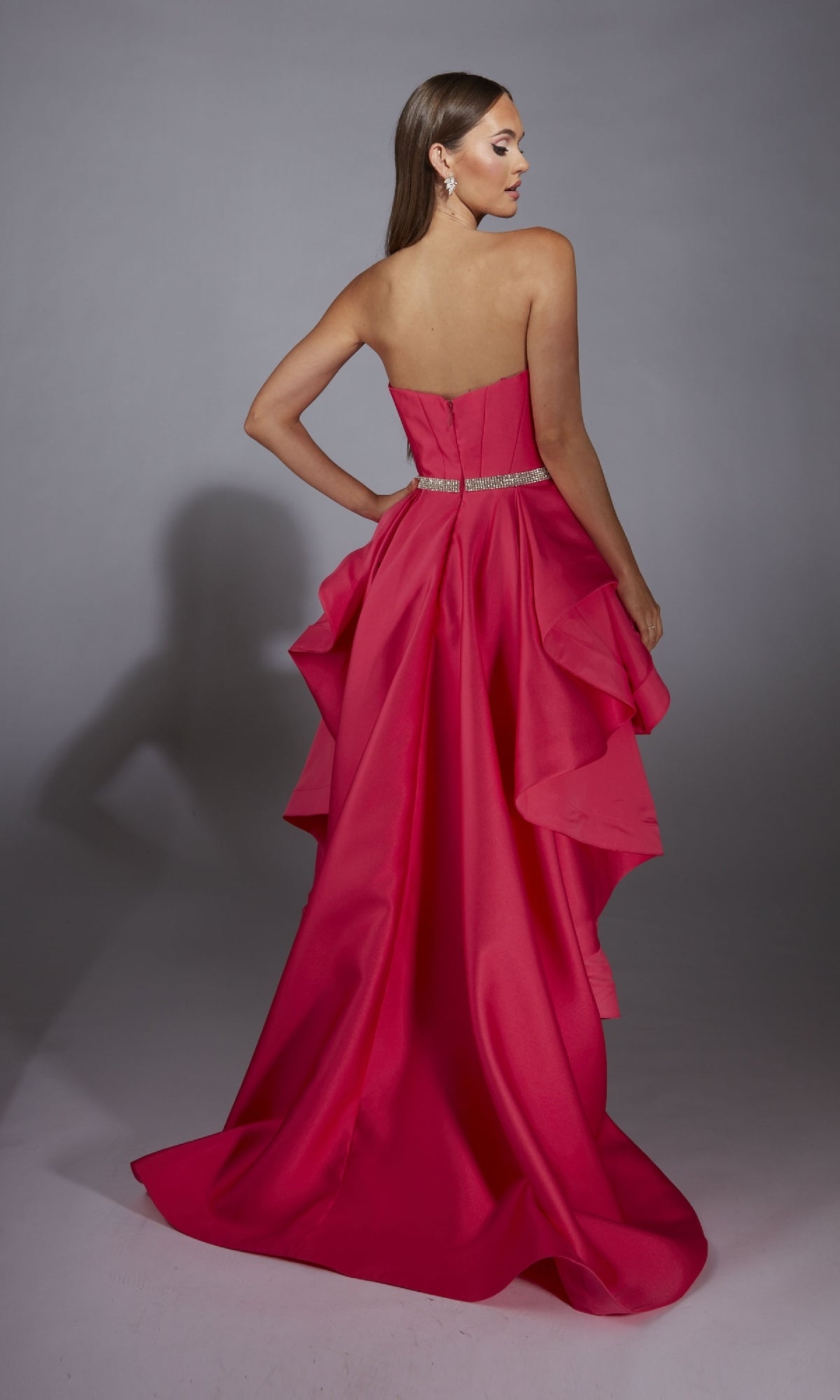 A woman in the Alyce 61934 high-low strapless prom dress in bright pink with a beaded waistline and layered ruffle back stands facing away against a plain gray background.