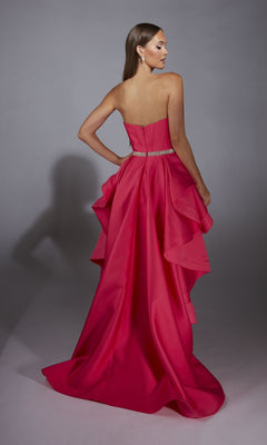 A woman in the Alyce 61934 high-low strapless prom dress in bright pink with a beaded waistline and layered ruffle back stands facing away against a plain gray background.