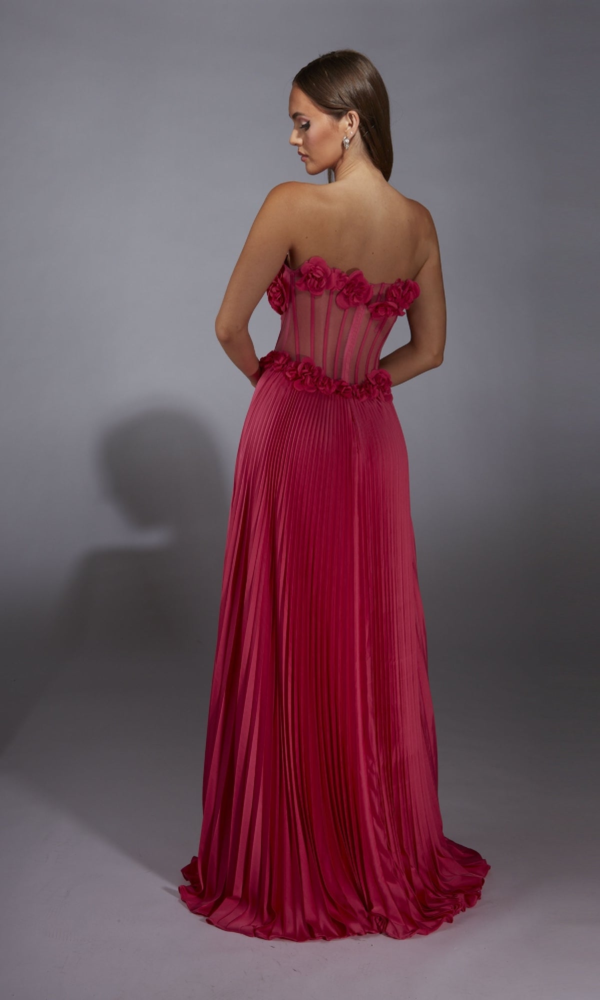 A woman faces away against a gray background wearing the Alyce 61935: a strapless, floor-length pink pleated prom dress with 3D floral details and a sheer corset bodice.