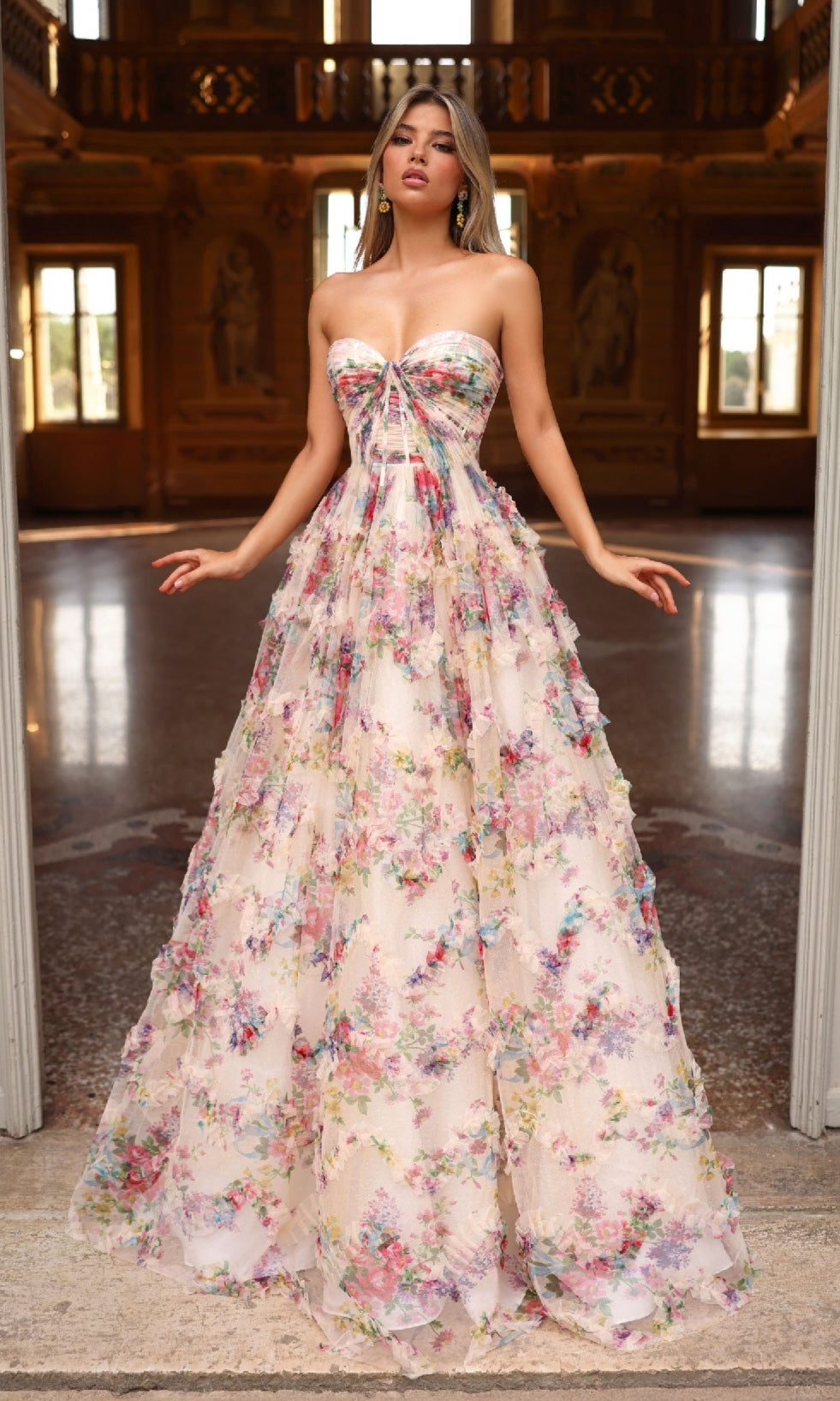 A woman stands in an ornate hall wearing the Alyce 61957 long prom dress, which features a strapless floral design with a delicate corset bodice and a flowing A-line skirt in soft pastel hues.