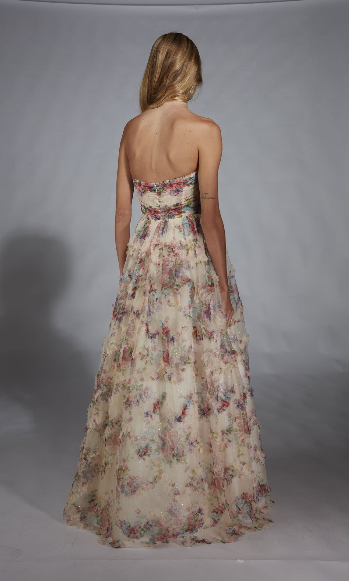 A woman with blonde hair faces away, modeling the Alyce 61957 long prom dress—strapless, with a floral corset bodice and elegant floor-length A-line skirt—against a plain gray background.
