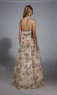 A woman with blonde hair faces away, modeling the Alyce 61957 long prom dress—strapless, with a floral corset bodice and elegant floor-length A-line skirt—against a plain gray background.