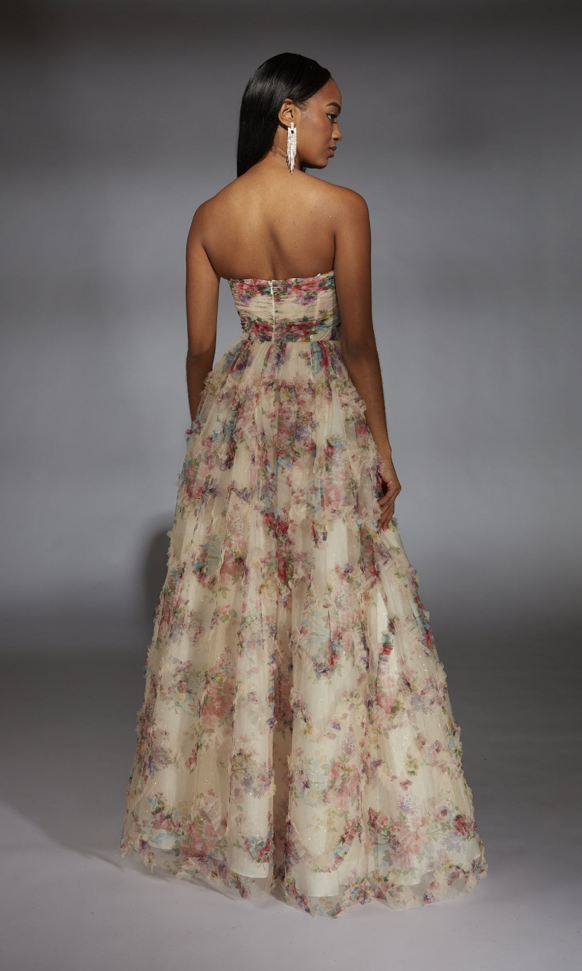 A woman faces away, modeling the Alyce 61957 long prom dress. The strapless floral gown features an A-line skirt with layered tulle, set against a plain studio backdrop.