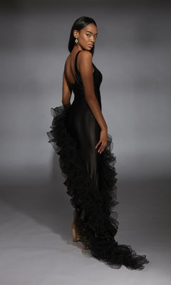 Wearing the Long Black Prom Dress: Alyce 61967 with ruffled trim and a high-low hem, a woman stands on a gray background, facing away and glancing over her shoulder.