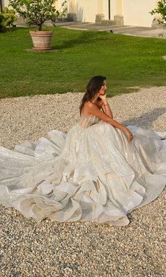 A woman wearing the Alyce 61971 long prom dress with a beaded corset bodice sits on a gravel path in an outdoor garden surrounded by grass and potted plants.