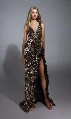 A person wears the Alyce 61975 Long Prom Dress—black with gold sequin floral patterns, deep V-neckline, thigh-high slit, ruffled trim, and strappy open back—standing against a plain gray background.