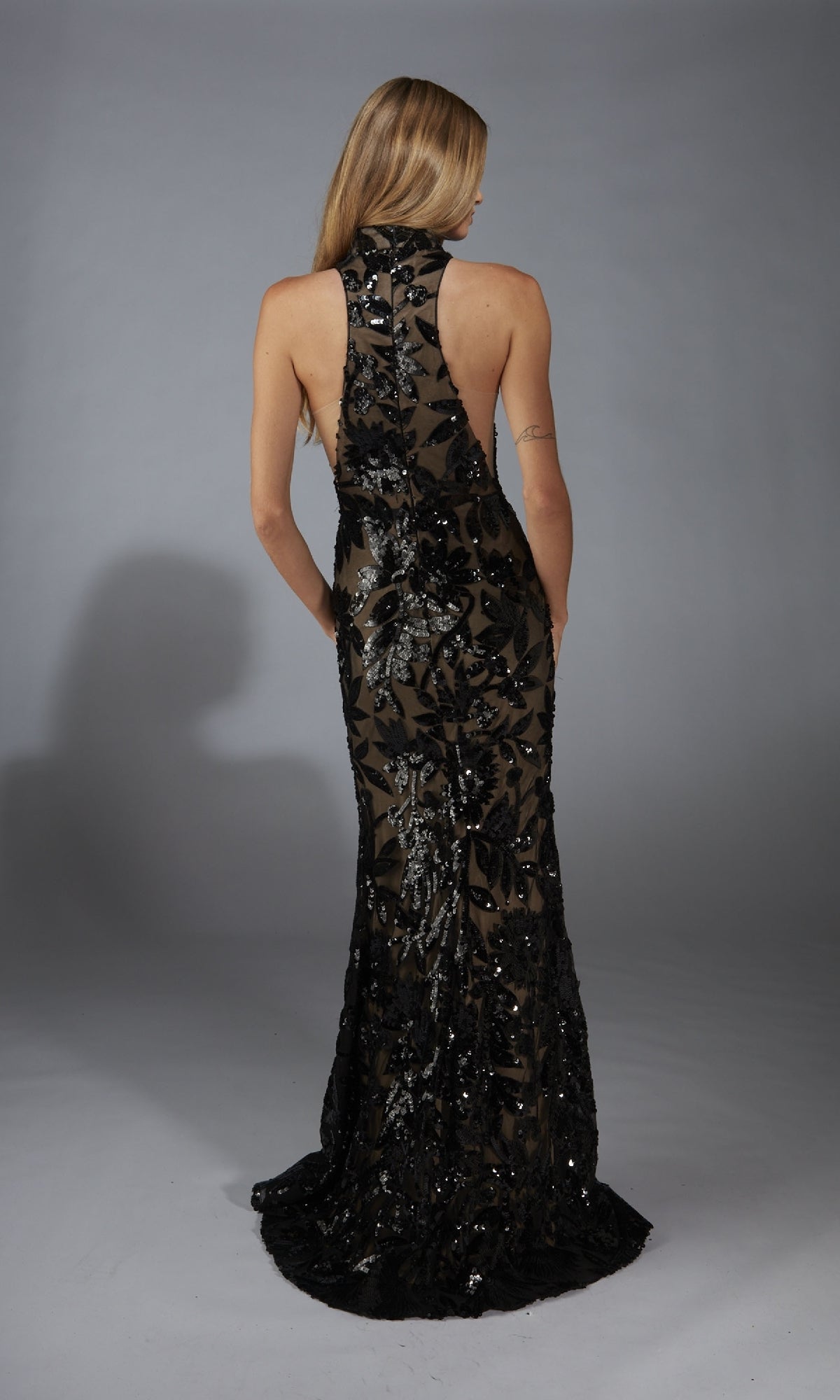 A woman with long blonde hair models the Alyce 61979, a sleeveless, floor-length black sequin prom dress with a sheer back, standing against a neutral studio backdrop.