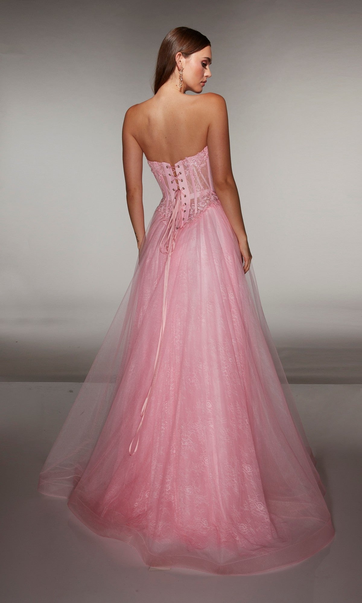 A woman wearing the Alyce 61984 Long Prom Dress with a strapless neckline and lace-up corset back stands facing away, her floor-length pink gown flowing elegantly against a plain background.