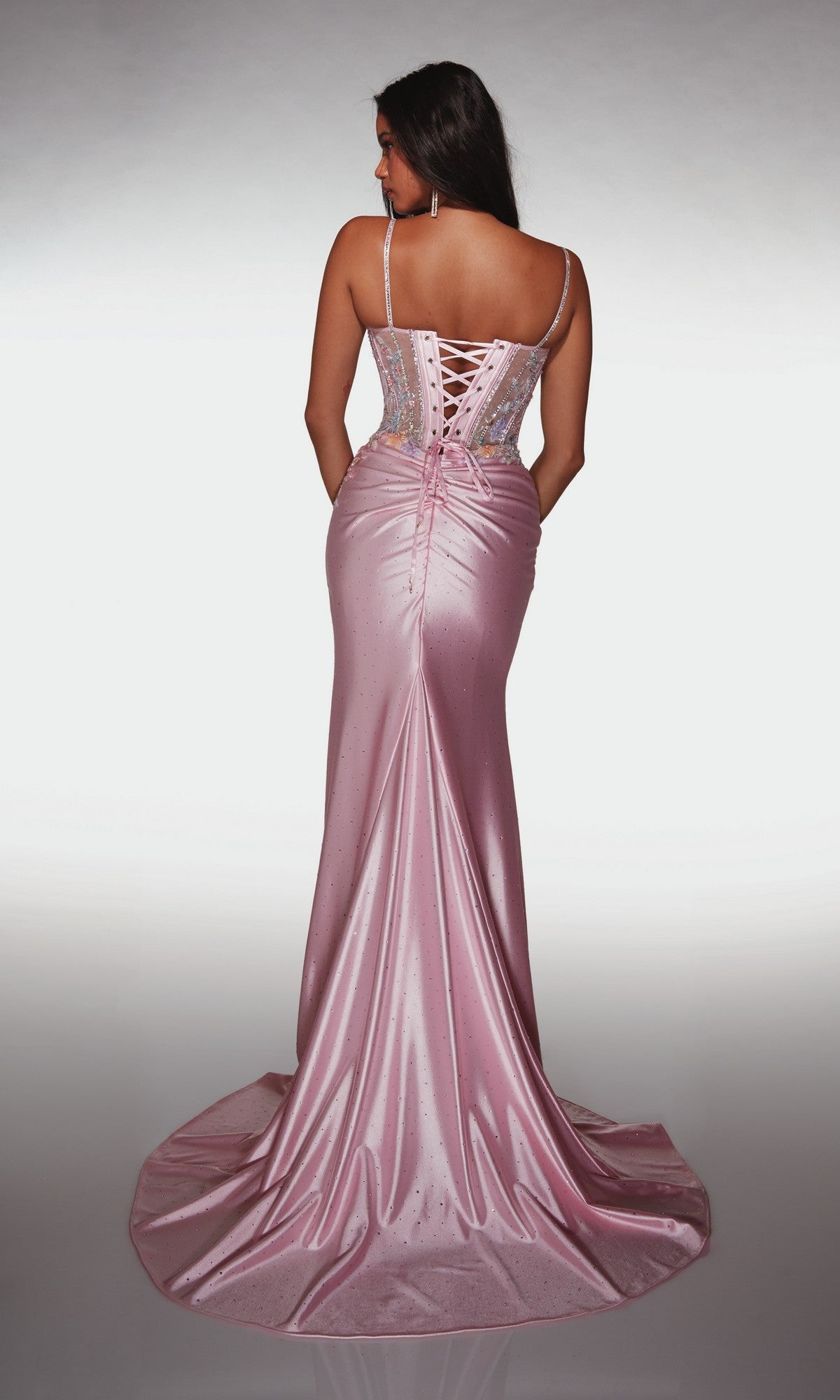 A woman wearing the Alyce 62022 long prom dress, featuring a shiny pink sleeveless design with a lace-up corset bodice and flowing train, stands facing away from the camera.