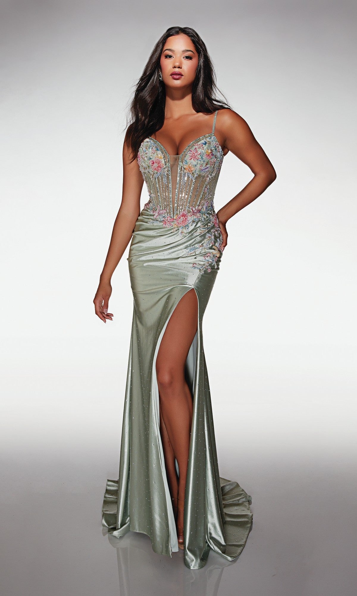 A woman stands against a plain background wearing the Alyce 62022 long prom dress—a light green, sleeveless gown with a corset bodice, high-leg slit, and floral embellishments.