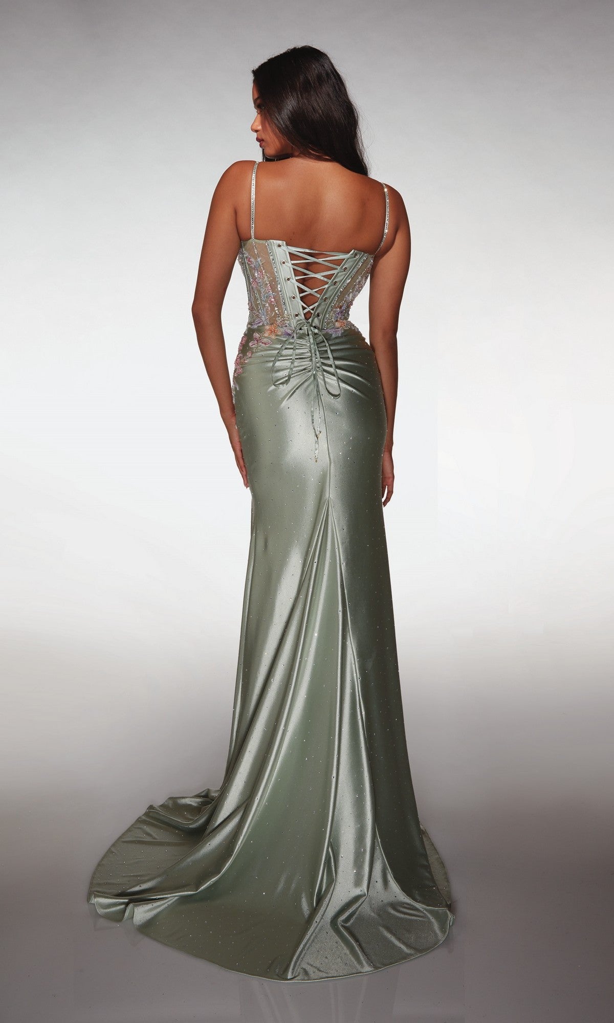 A woman stands facing away, wearing the Alyce 62022 long prom dress—a shimmery light green, sleeveless gown with a corset bodice, lace-up back, and flowing train.