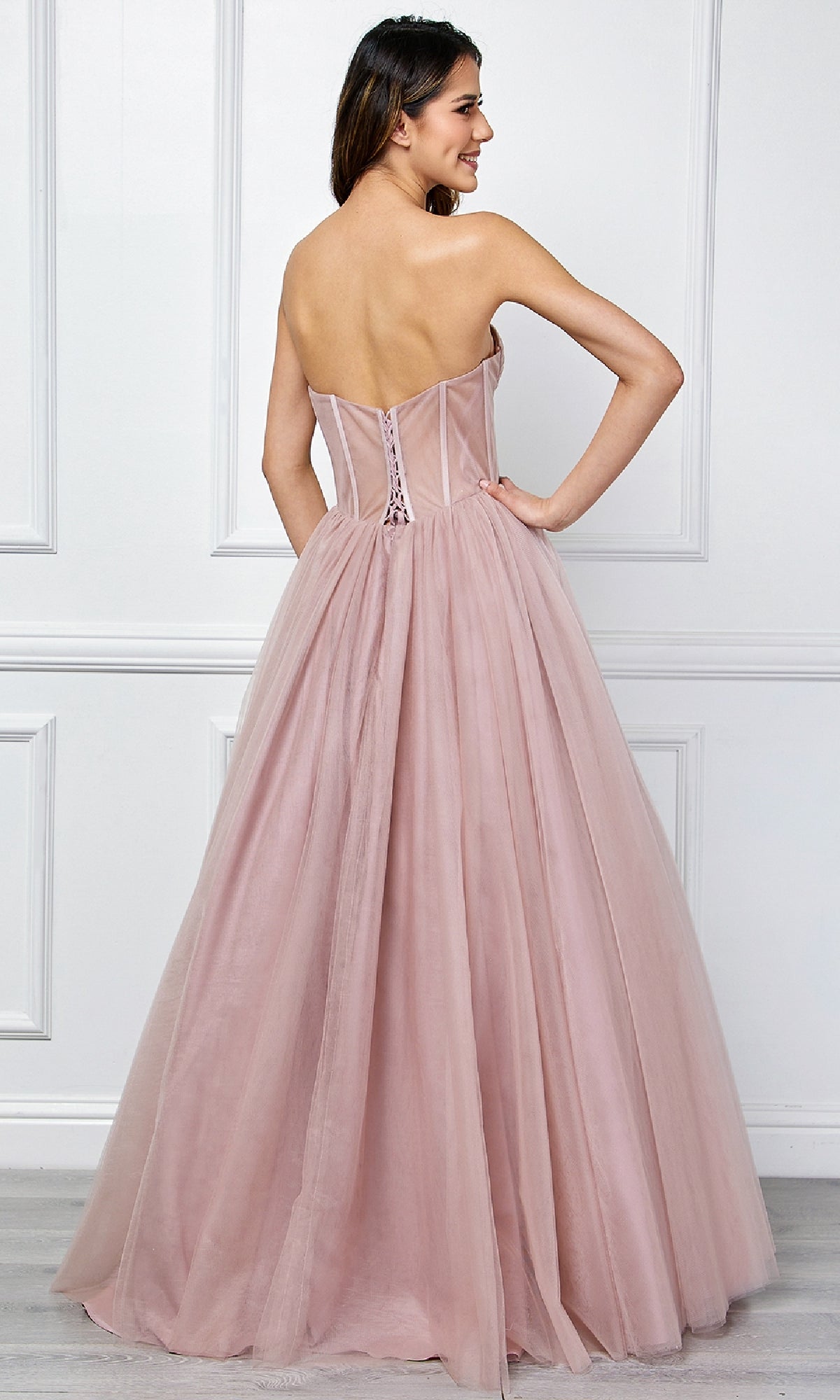 A woman faces away from the camera in a blush pink Amelia Couture 6202 strapless corset prom ball gown, featuring a full tulle skirt, standing before a white paneled wall—an ethereal look.