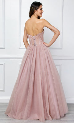 A woman faces away from the camera in a blush pink Amelia Couture 6202 strapless corset prom ball gown, featuring a full tulle skirt, standing before a white paneled wall—an ethereal look.