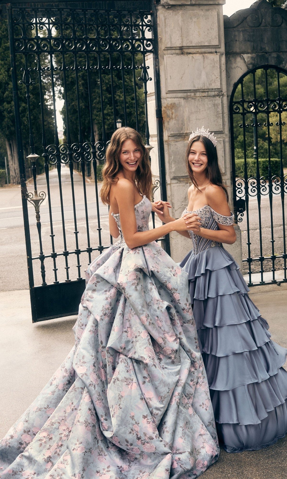 Two young women stand smiling before a wrought iron gate; one dazzles in the Alyce 62035 long prom dress, featuring ruffles, a beaded corset bodice, and a tiara, while the other wears a floral ballgown.