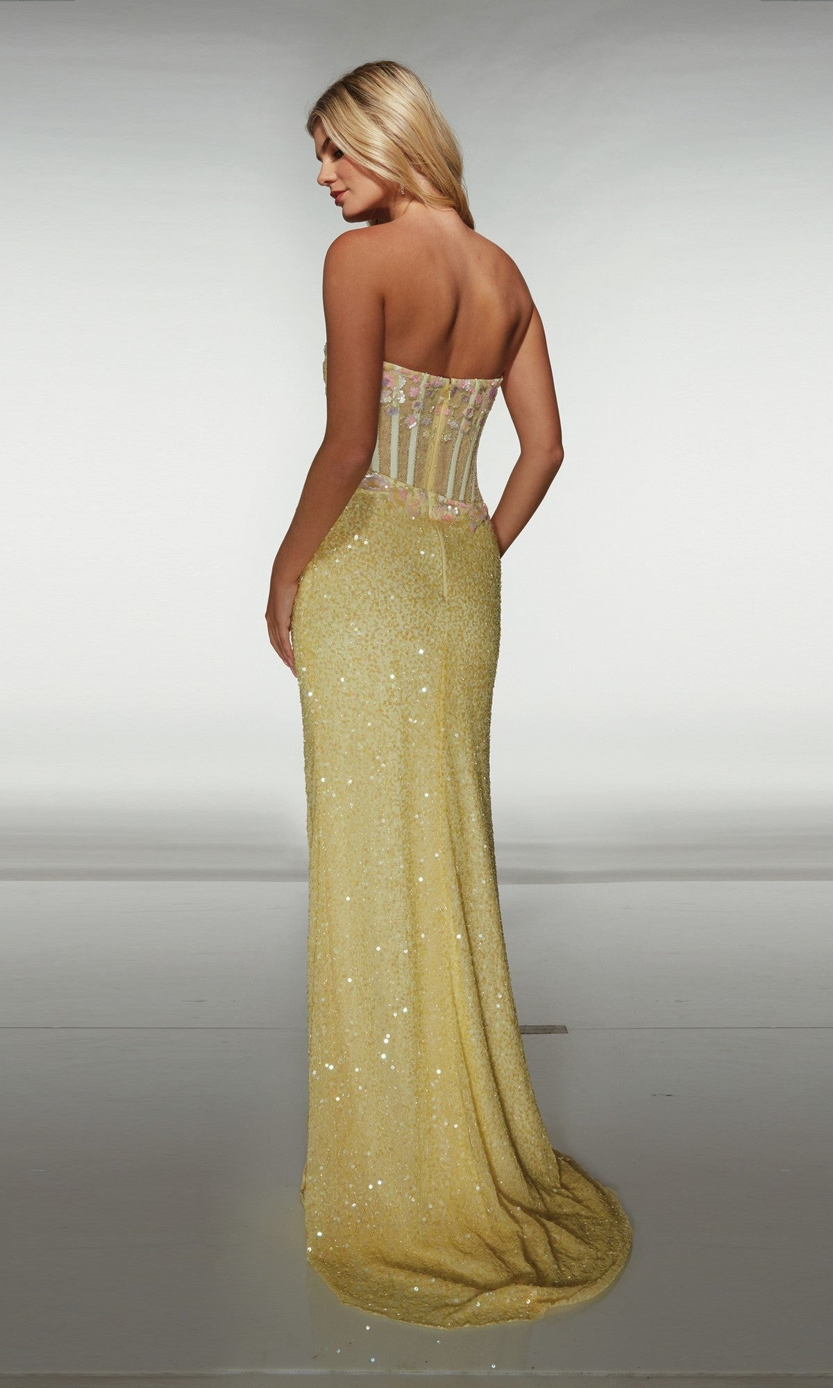 A woman faces away from the camera in an Alyce 62047 long prom dress, featuring a strapless yellow floor-length design with a fitted bodice, sheer back panel, and sequin wrap-style skirt against a plain background.