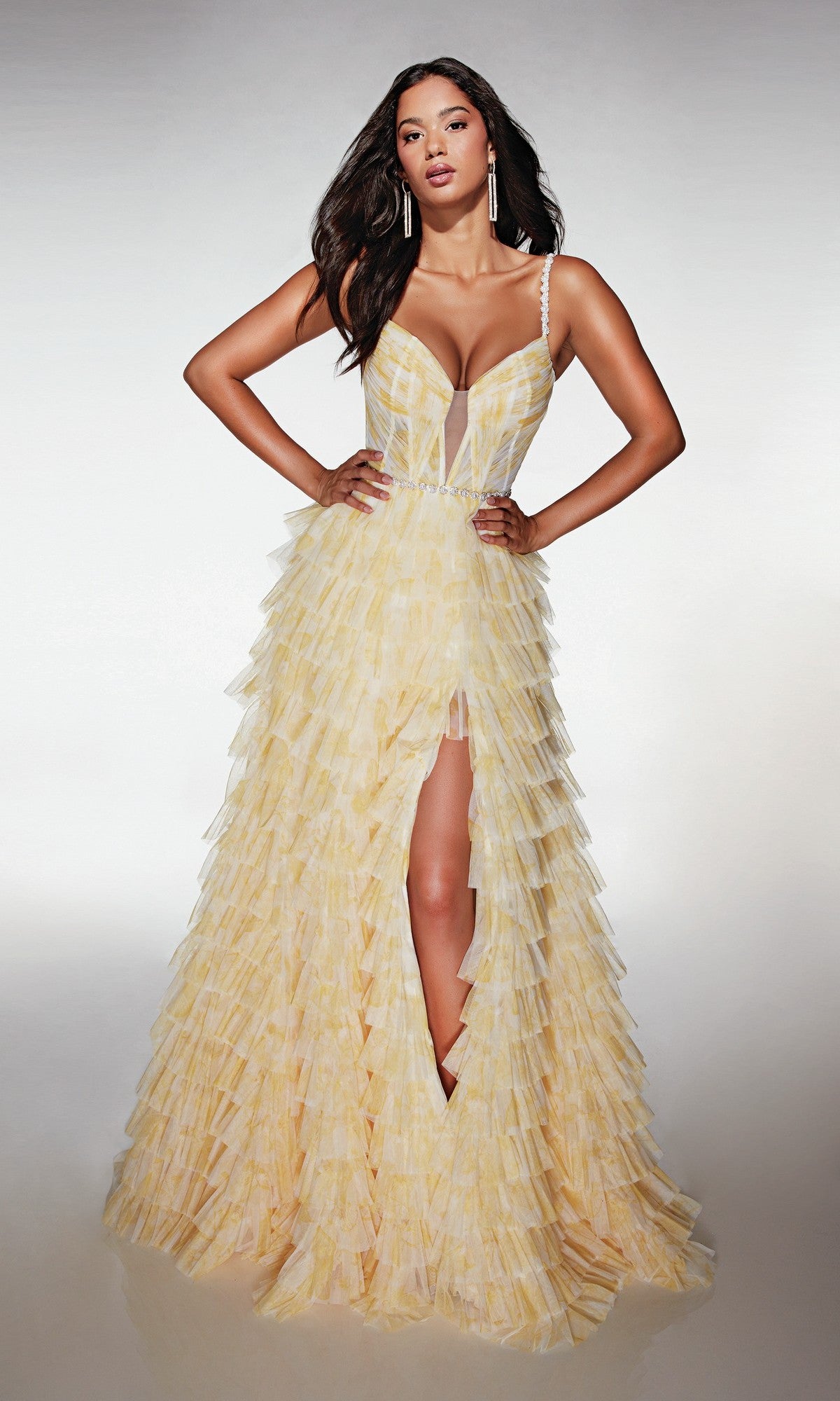 A woman poses with hands on hips in the Alyce 62052 long prom dress in Buttercup—a sleeveless gown with a deep neckline, high slit, and tiered ruffle skirt—set against a plain background.