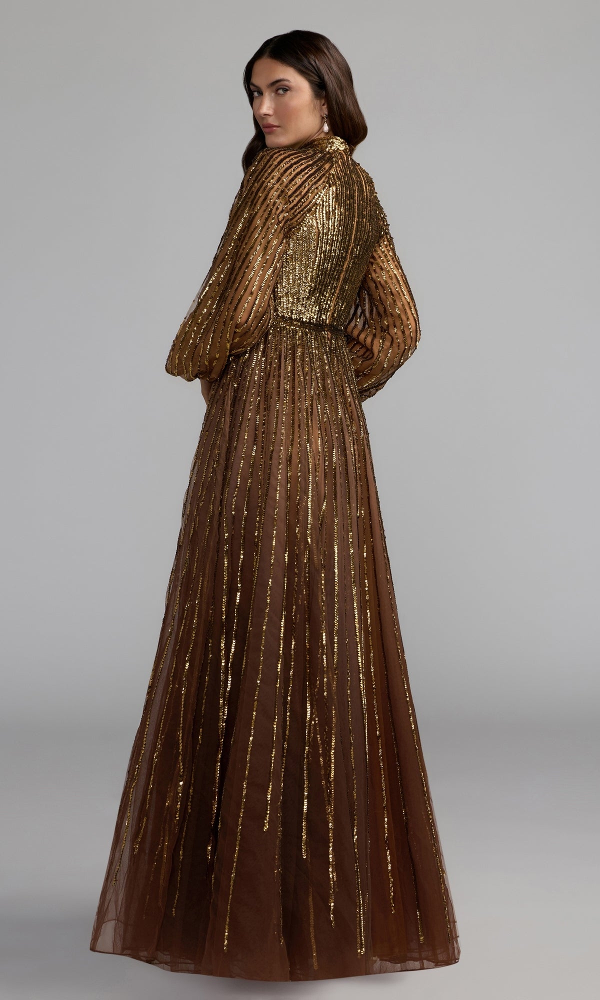 A woman stands facing away, looking over her shoulder against a plain gray background, wearing the refined and elegant Blue Ombre Sequin Formal Gown (Mac Duggal 6206).