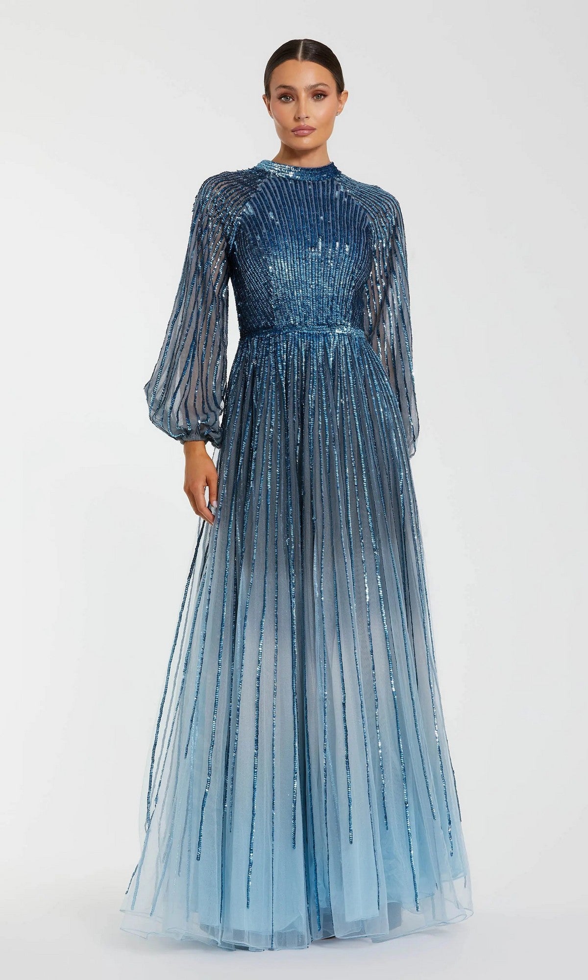 A woman wears the Mac Duggal 6206 long formal dress, a blue ombre sequin gown with sheer long sleeves and vertical sequin detailing, standing against a plain white background.