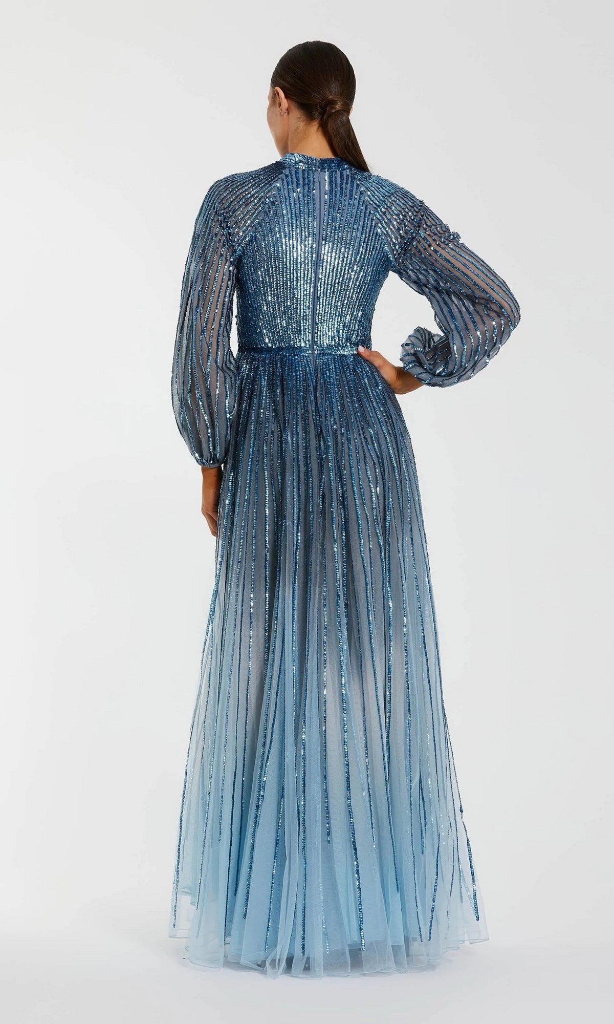 A woman faces away, wearing the Mac Duggal 6206—a long formal dress in blue ombre sequins, featuring sheer striped sleeves and a flowing skirt. Her hair is tied back in a low ponytail.