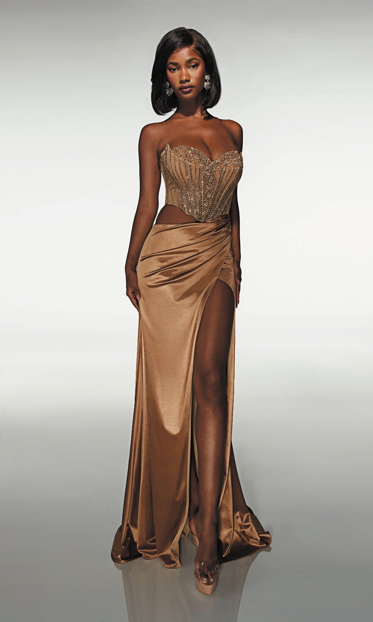 A woman wears the Alyce 62087 long gold prom dress with a strapless beaded corset bodice and leg slit, paired with clear heels, posing against a plain light background.
