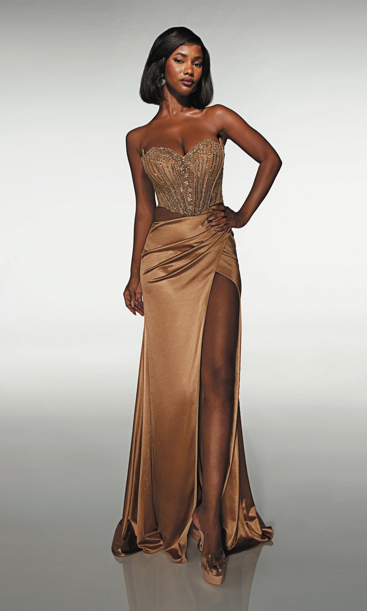 A woman models the Alyce 62087 long prom dress—a strapless gold satin gown featuring a beaded corset and high slit—against a plain background.