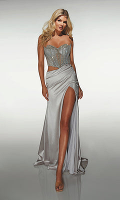 A woman models the Alyce 62087 long prom dress, featuring a silver hue, beaded corset bodice, and daring leg slit, posed against a plain light background.