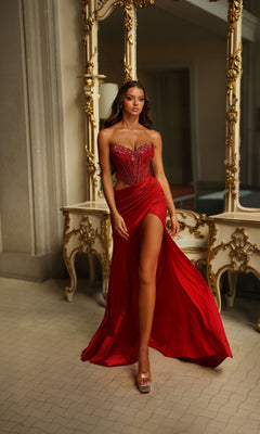 Wearing the Alyce 62087 long prom dress featuring a red hue, sweetheart neckline, and high slit, a woman stands before an ornate mirror and table in an elegant interior.