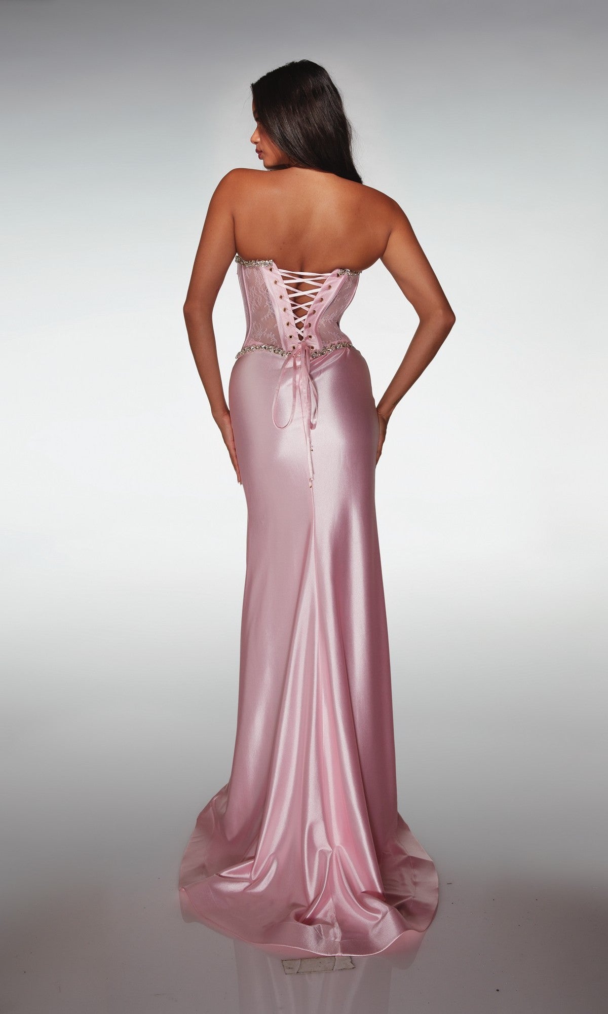 A woman stands with her back to the camera, wearing the Alyce 62089— a long prom dress in pink satin with a strapless lace corset bodice and elegant lace-up back detailing.