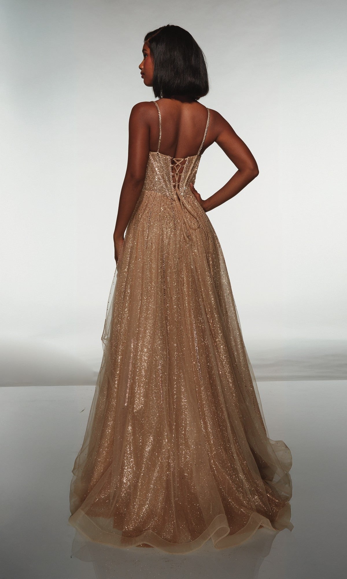 A woman faces away, wearing the Alyce 62093 long prom dress—a floor-length gold glitter tulle gown with thin straps and a lace-up back—set against a plain light background.