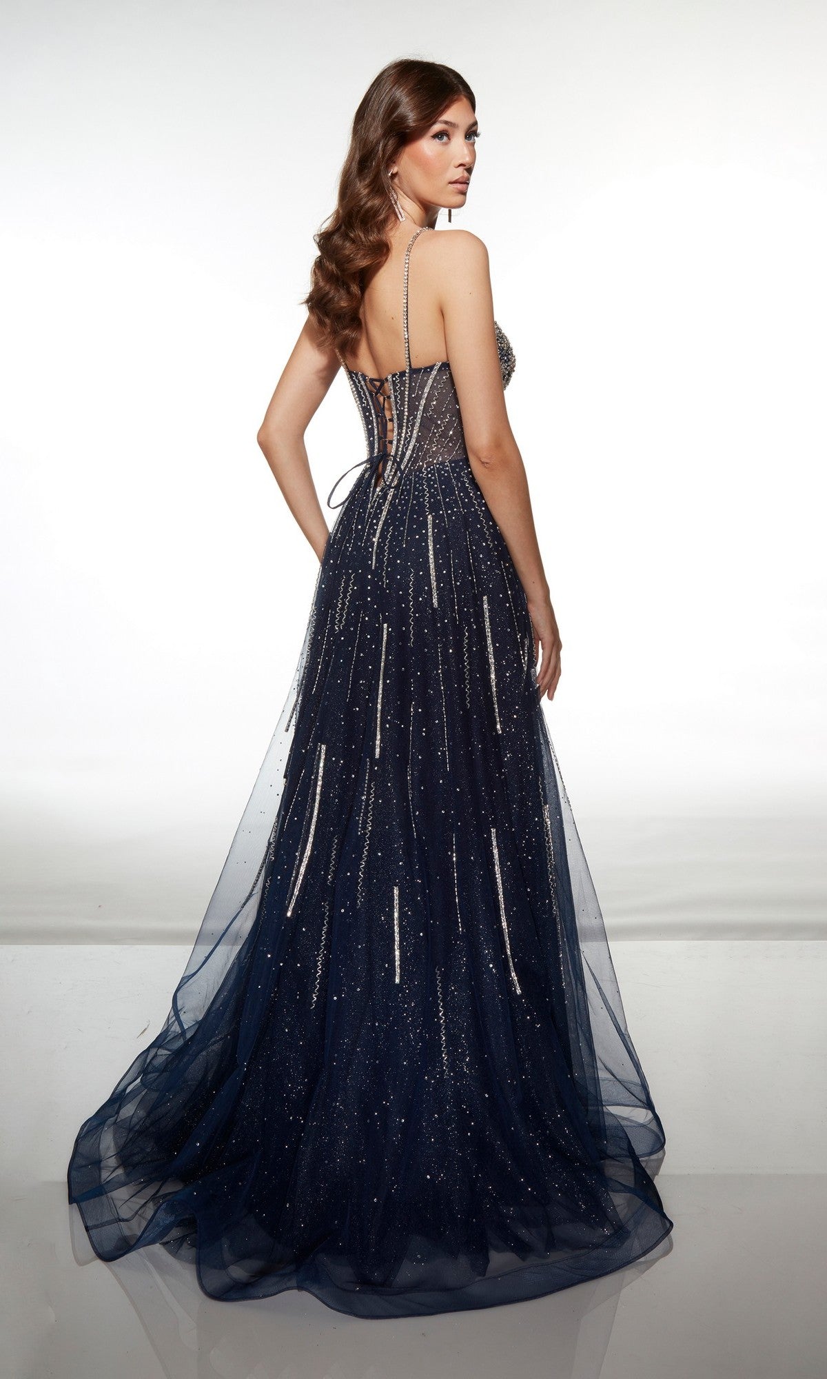 A woman stands facing away, wearing the Alyce 62093 long prom dress—a dark blue glitter tulle gown with thin straps, lace-up back, and a floor-length sparkling finish.