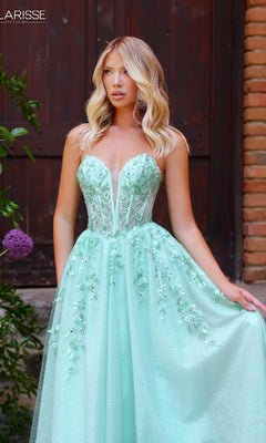 A woman in the Clarisse Designs 62102 prom ball gown—strapless, mint green glitter tulle with beaded floral lace—stands outdoors by a wooden door and brick wall, holding her dress.