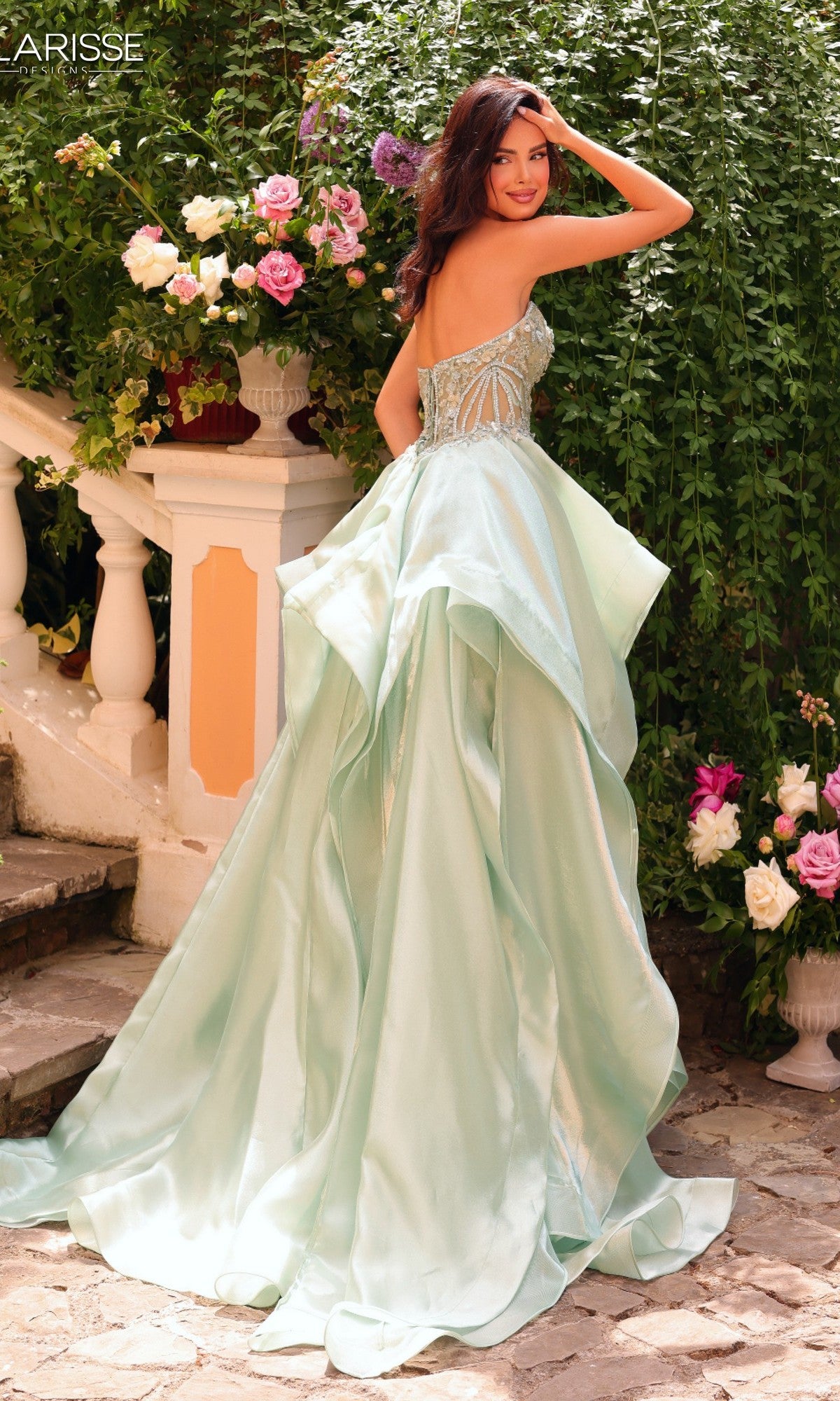 A woman in the Clarisse Designs 62131 Prom Ball Gown, featuring a strapless satin silhouette with a beaded corset bodice, stands outdoors near steps and flowers, glancing back over her shoulder.