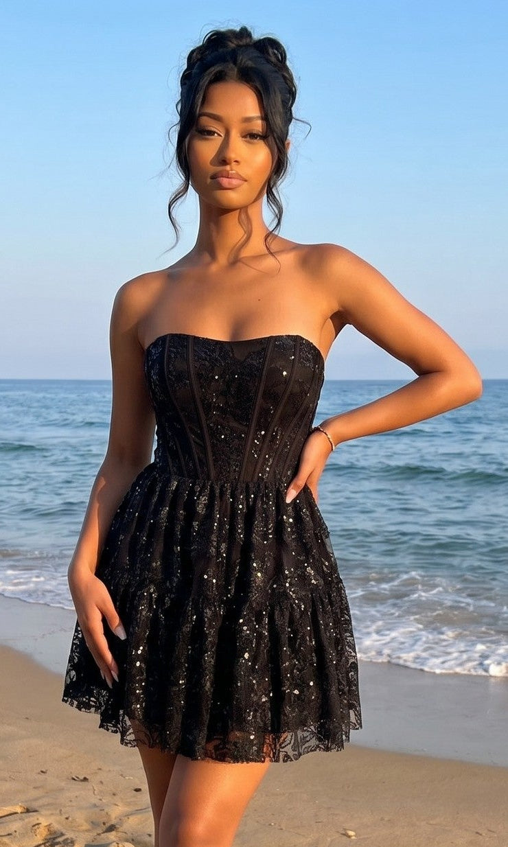 A woman wearing the Strapless Mini Hoco Dress: Blondie Nites 6213BN in black stands on a sandy beach with the ocean and blue sky in the background.