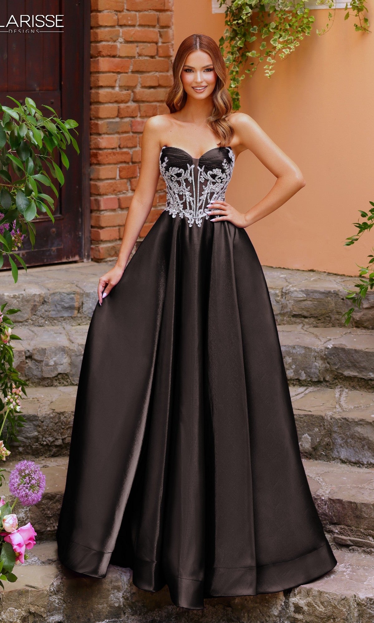 A woman in a black Clarisse Designs 62140 prom ball gown with a strapless beaded corset bodice and silver embroidery stands on stone steps before a brick wall and plants.