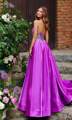 A woman wearing the Clarisse Designs 62140 prom ball gown, featuring a strapless beaded corset bodice and floor-length bright purple satin, stands outdoors by flowers and stone steps, glancing back over her shoulder.