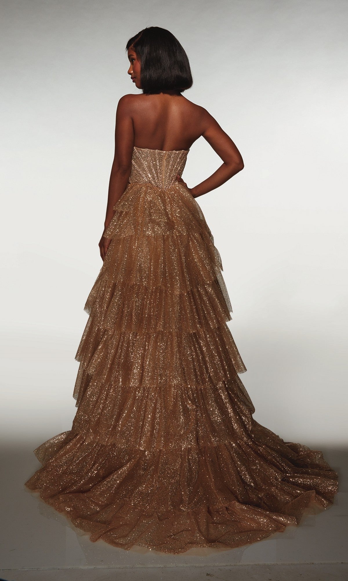 A woman wearing the Alyce 62144 long formal dress featuring a gold, glittery tiered ruffle skirt stands with one hand on her hip, facing away against a plain background.